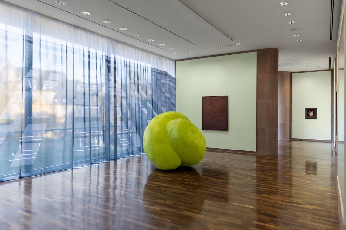 In an exhibition room of the Kunsthalle. In front of the wall-high window is a yellow-green, roundish object, about 1.60 meters high. In front of the window a curtain with an architectural photograph printed on it. Behind it, paintings on two walls.