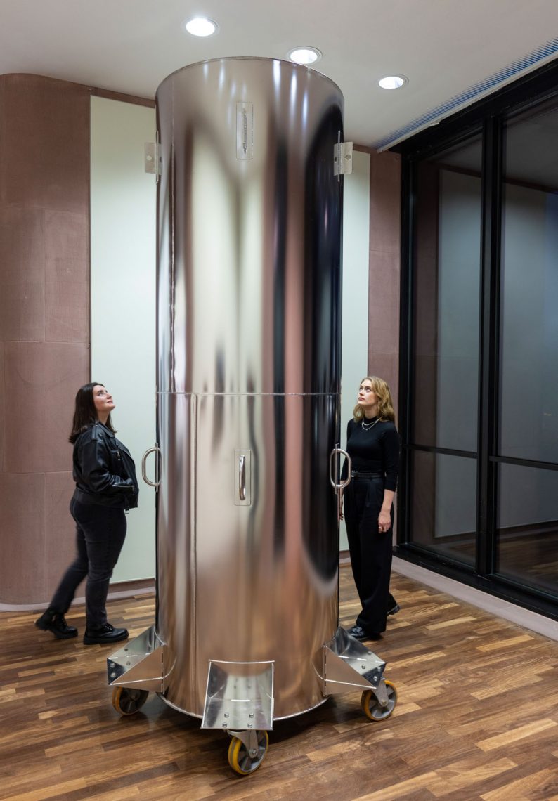 In an exhibition room, an almost floor-to-ceiling shiny silver metal cylinder on castors, vertical. Behind it are two women, dressed in dark clothes, looking up at the cylinder.