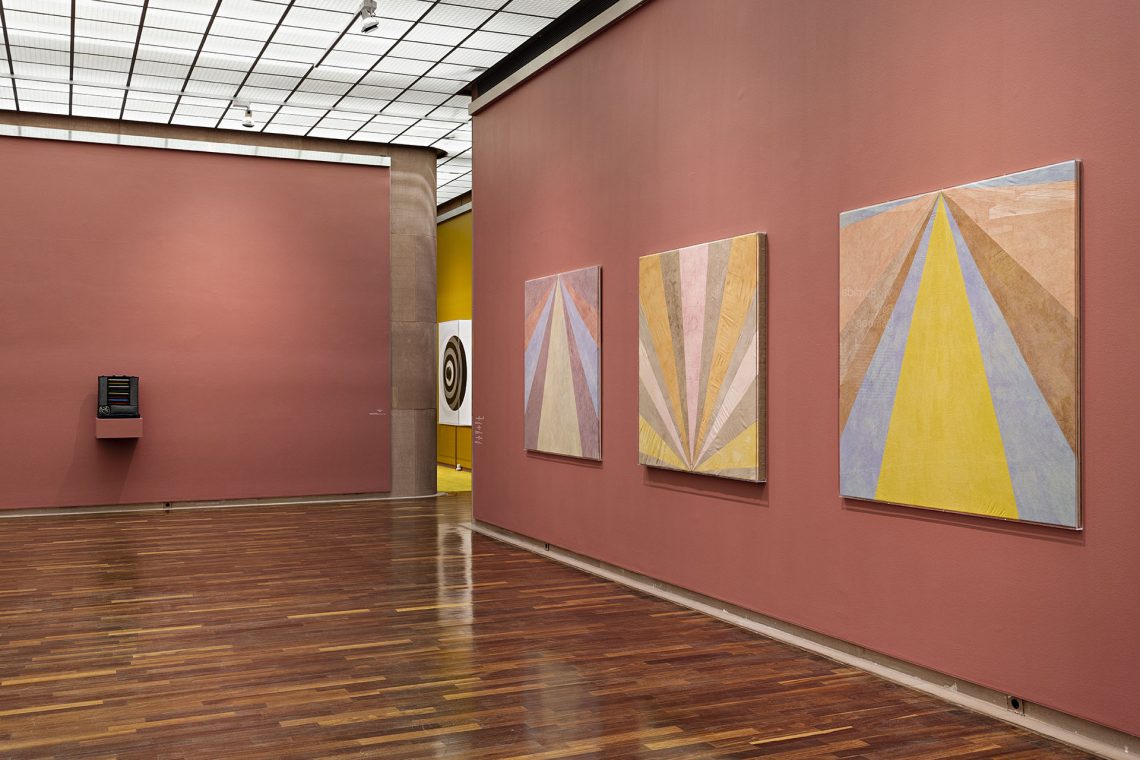 Mehrere Wände. Rechts auf hellroter Wand drei quadratische Werke mit strahlenartig von einem Punkt am oberen oder unteren Rand ausgehenden Dreiecken in grau, dunkelgelb, orange, hellrot und zartrosa.