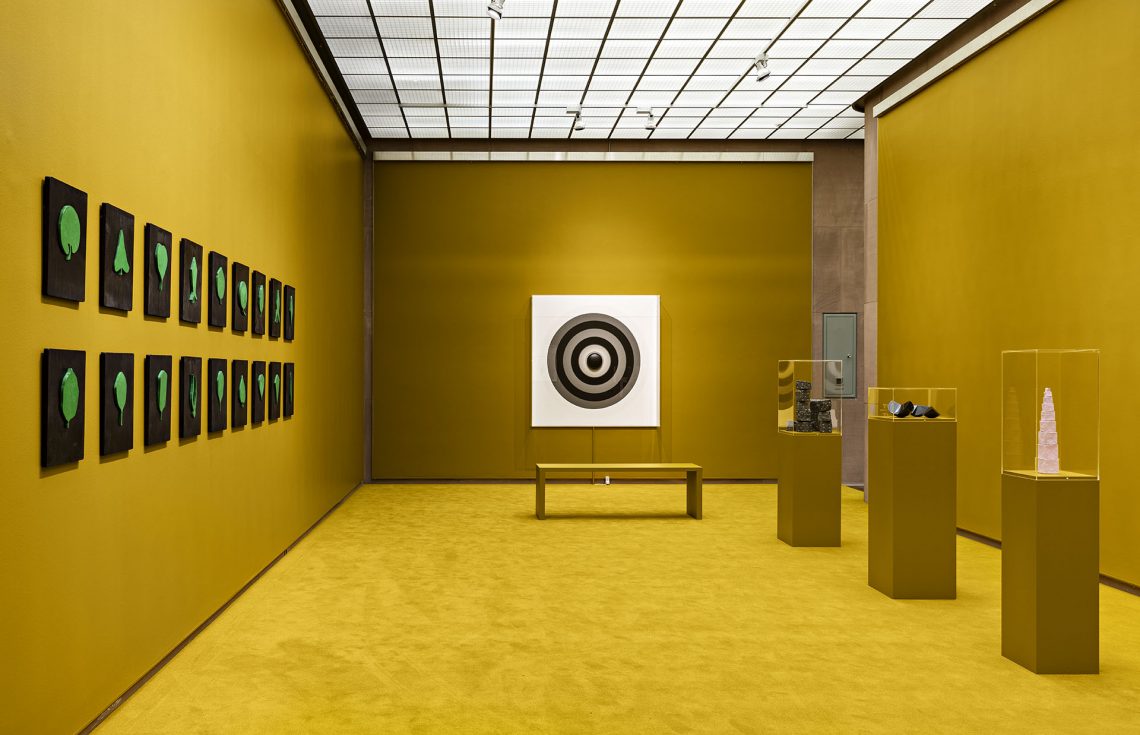 Ein goldfarbener Raum mit einer Kreisform auf einem weißen Quadrat an der hinteren Wand. Links grüne Blattformen auf schwarzen Rechtecken, rechts kleinere Kunstobjekte auf Sockeln. Vor dem Quadrat hinten eine Sitzbank ohne Lehne.