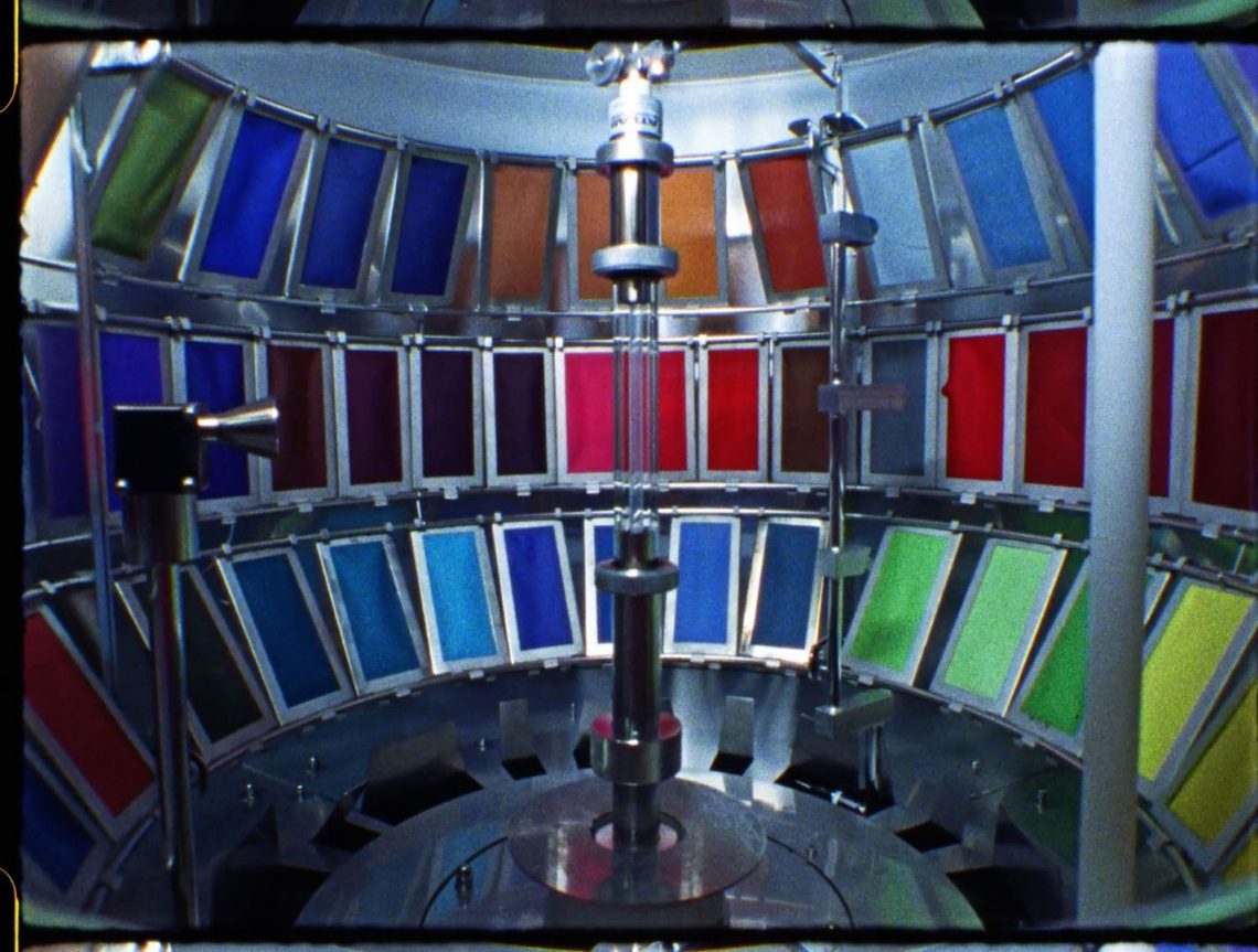 Zu sehen sind 3 Reihen von Stoffproben in vielen verschiedenen Farben, die in einem Apparat um eine neonlampenartige Röhre aufgespannt sind.