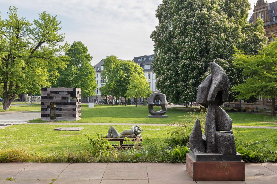 Sculptures in a park. In front on the right a kind of figure made of simple shapes, behind it a reclining female figure, behind it a cube-shaped small wooden house, further away a large oval bronze sculpture. Trees all around, in the distance a multi-story apartment building.