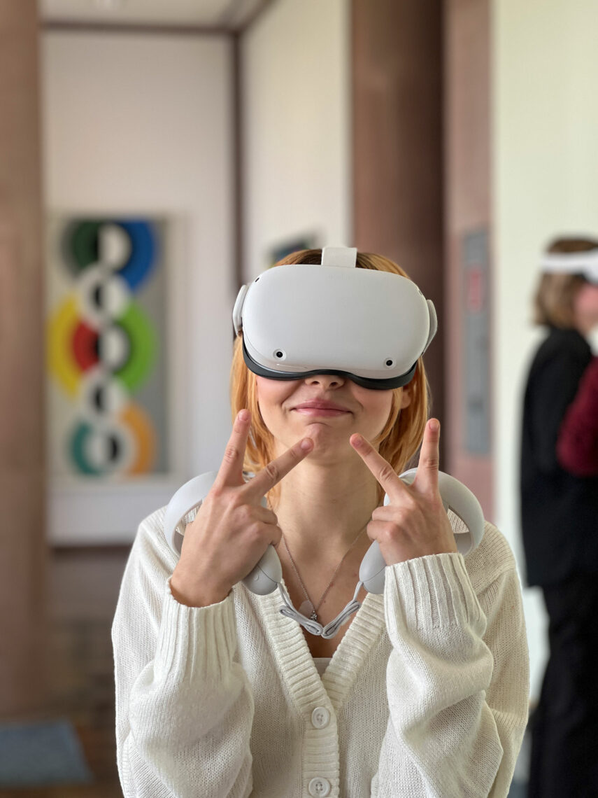 Eine junge Frau in einem Kunstmuseum. Sie trägt ein Virtual Reality Headset und schaut in unsere Richtung. Sie hat gute Laune.