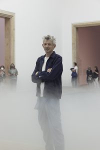 Middle aged white man with curly gray hair. He stands in an open shirt and t-shirt with folded arms in a showroom.