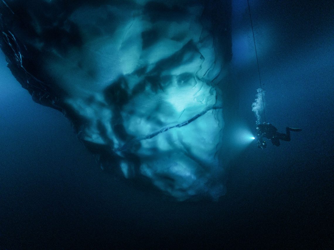 Ein Taucher, der im tiefblauen Wasser um einen Eisberg herum taucht. Das Tageslicht und das seiner Taschenlampe reflektieren im und am massiven Eisberg.