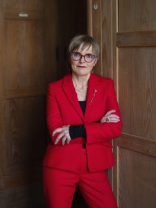 Middle aged white woman with short light blond hair and big red, glasses. She stands with folded arms in a light red suit.