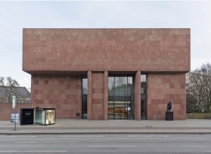 The façade of a rectangular building made of red sandstone. The entrance with floor-to-ceiling windows can be seen from the street. In front of it on the left is a small bus stop shelter, on the right a seated bronze figure on a pedestal.