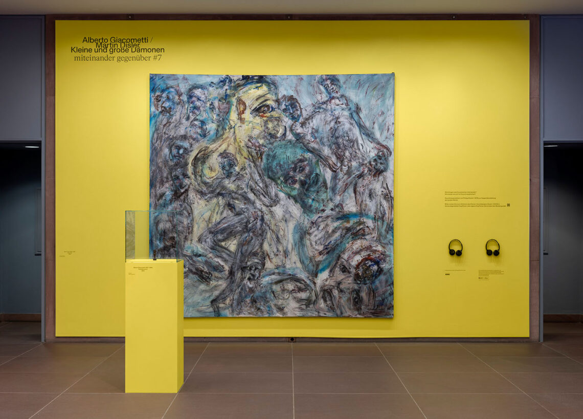 Eine gelb gestrichene Wand in der Kunsthalle Bielefeld, darauf ein quadratisches Gemälde von Martin Disler mit wild wirbelnden Figuren. Rechts davon zwei Kopfhörer. Davor im Raum ein etwa 1,30 Meter hoher Sockel mit einer sehr kleinen Skulptur von Alberto Giacometti.