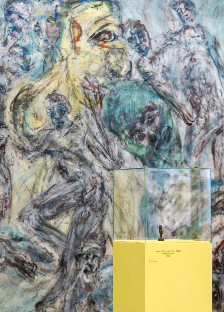 Ein Gemälde von Martin Disler mit wild wirbelnden Figuren. Davor im Raum ein Sockel mit einer sehr kleinen Skulptur von Alberto Giacometti.