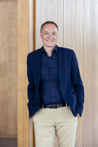 A white man stands leaning against a wooden wall with his hands in his pockets. He has short hair, wears a blue jacket and shirt and beige pants.