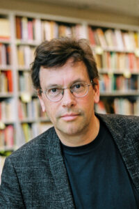 A middle-aged white man with glasses and short dark brown hair. He wears a dimly checkered black sacko over a black T-shirt. A filled bookshelf is blurred in the background.