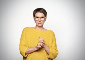 A middle aged white woman against a white background. She wears yellow knitted sweater, round glasses with wide yellow frame and short dark brown hair.