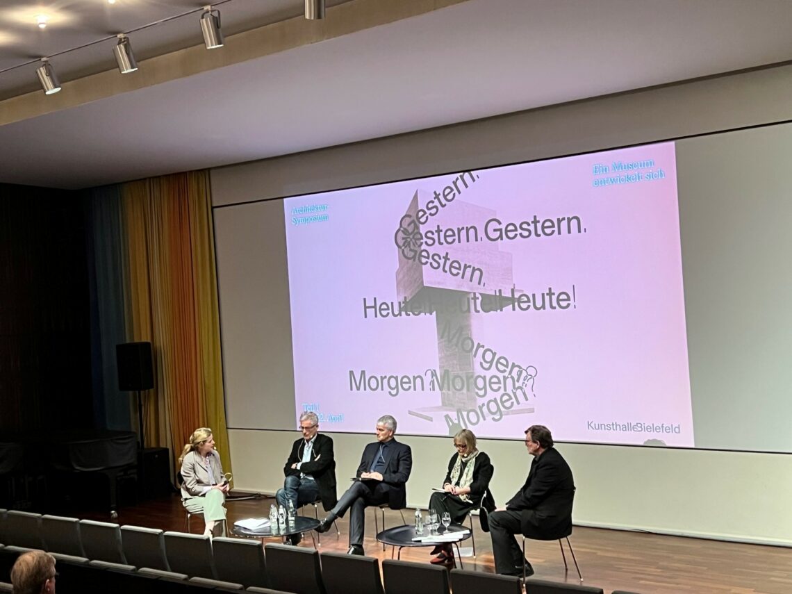 Three white men and two white women sit with chairs on a stage. A poster for the architecture symposium is beamed onto the wall Behind the panel.