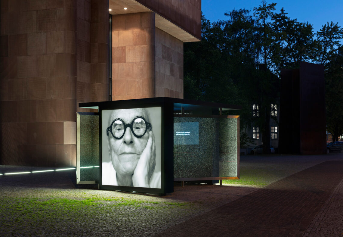 The illuminated bus stop shelter in front of the Kunsthalle Bielefeld at night. At its head is a black and white portrait photo of Philip Johnson wearing black, round glasses. The rest of the glazing is intentionally shattered.