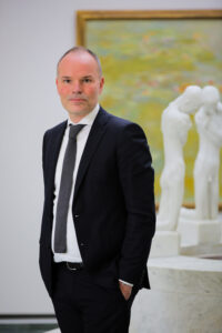 A gentleman in a black suit, white shirt and dark tie stands in a white room with his hands in his pockets. Behind him, two white statues and a painting are blurred.