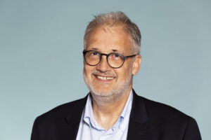 Portrait photo of a white, middle-aged man. He is laughing and has short white hair and beard, wears round glasses with black frames, blue and white checkered shirt and dark blue jacket.