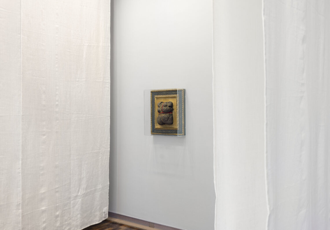 A brown lump of cloth wrapped with twine is mounted in an old picture frame. The background is golden, the wide border blue and gold, with a fine pattern. A plexiglass hood serves to protect the work. On both sides, white fabric panels hang from the ceiling.