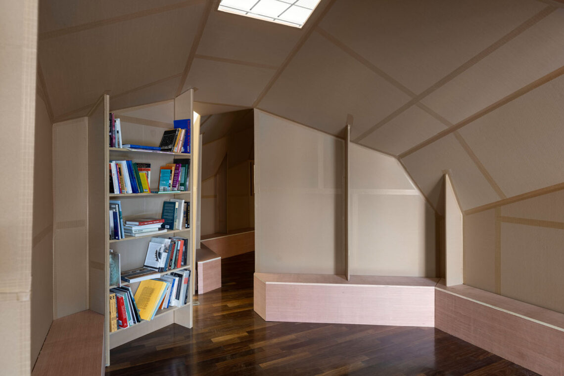 Blick in ein igluartiges Papphaus mit Sitzbänken an den Seiten. Links ist ein raumhohes Regal mit Büchern darin.