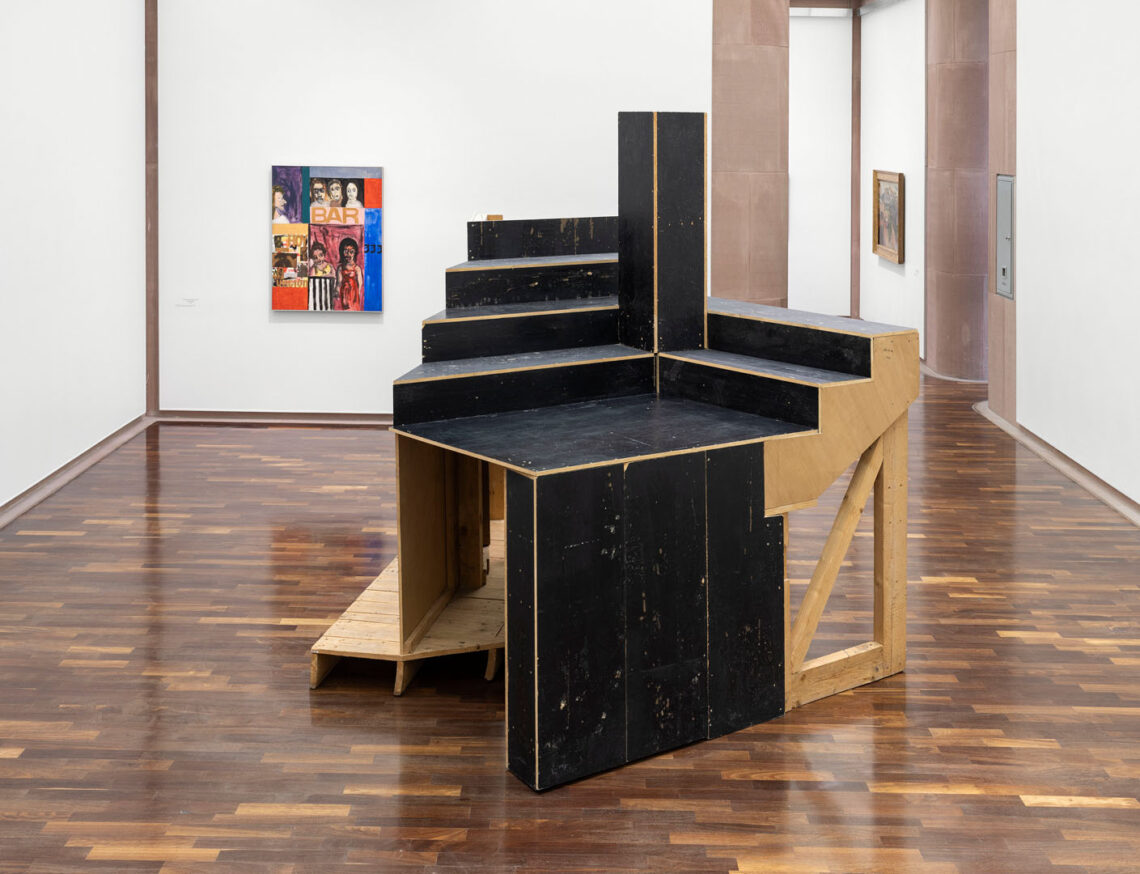 In einem Ausstellungsraum der Kunsthalle steht eine große schwarze und naturfarbene Holzstruktur, die an eine Treppe oder Tribüne erinnert. Wie ein Ausschnitt daraus. Hinten links an der Wand ein Gemälde mit Frauengesichtern und dem Wort "Bar".