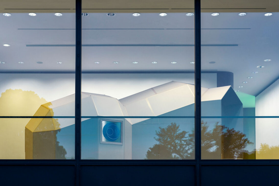 Blick von außen in ein Fenster der Kunsthalle, hinter dem ein igluartiges Haus zu sehen ist. In der großen Fensterfront spiegelt sich der Himmel, wodurch manche Teile des Fotos blau, manche gelb eingefärbt sind.
