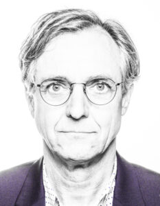 A man between 50 and 60 years old with a round face and glasses with thin metal frames. He wears his hair parted to the side, the hairline is slightly higher. He wears a fine light shirt with dark spots and a purple jacket over it.