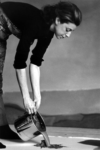 Schwarz-weiß Fotografie einer Frau, die Farbe aus einem Topf auf den Boden gießt.