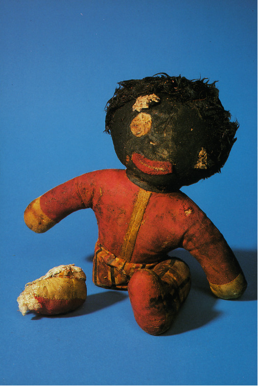 An old rag doll with red body and black head and emphasized wide lips. She looks very battered. One eye is missing, one leg too.