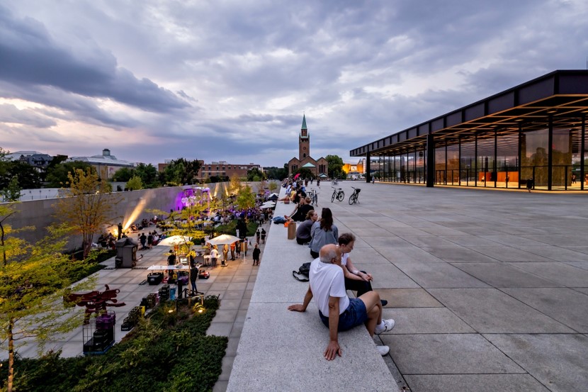 Evening twilight. On the right is a modern building with large windows. Bordering it on the left is a paved area with people sitting along its edge. You can look down to another paved area on the left. There are many people, tables, pavilions, surrounded by bushes and small trees.