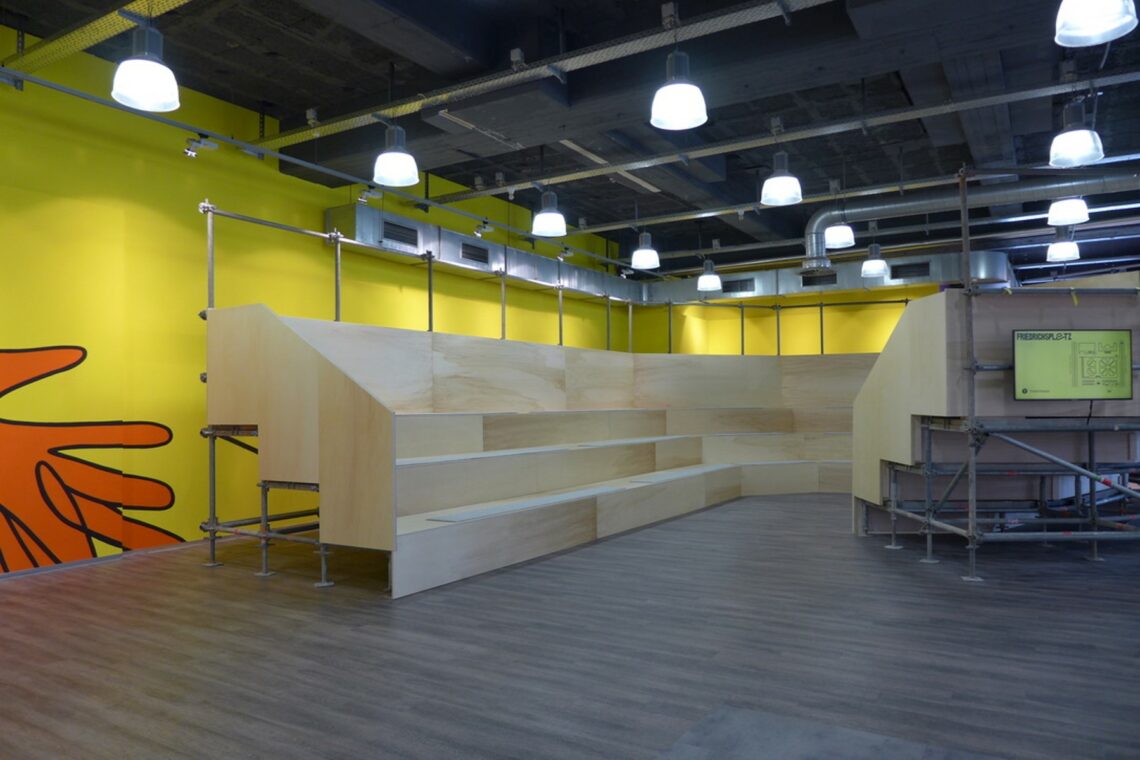 In einem gelb gestrichenen hallenartigen Raum steht eine große Sitzmöglichkeit. Die treppenartige Konstruktion ist aus Stangen von Baugerüsten und Holzplatten gebaut.