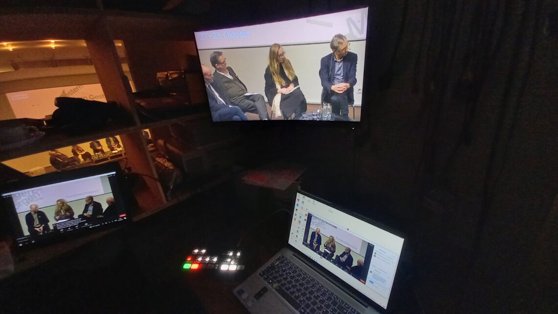 Arranged in a triangle, there is a large TV screen at the top, an open laptop at the bottom right and a tablet at the bottom left. All three screens show people sitting on chairs and talking. If you look at all the screens together, they show eight different people, but from different perspectives on each screen.