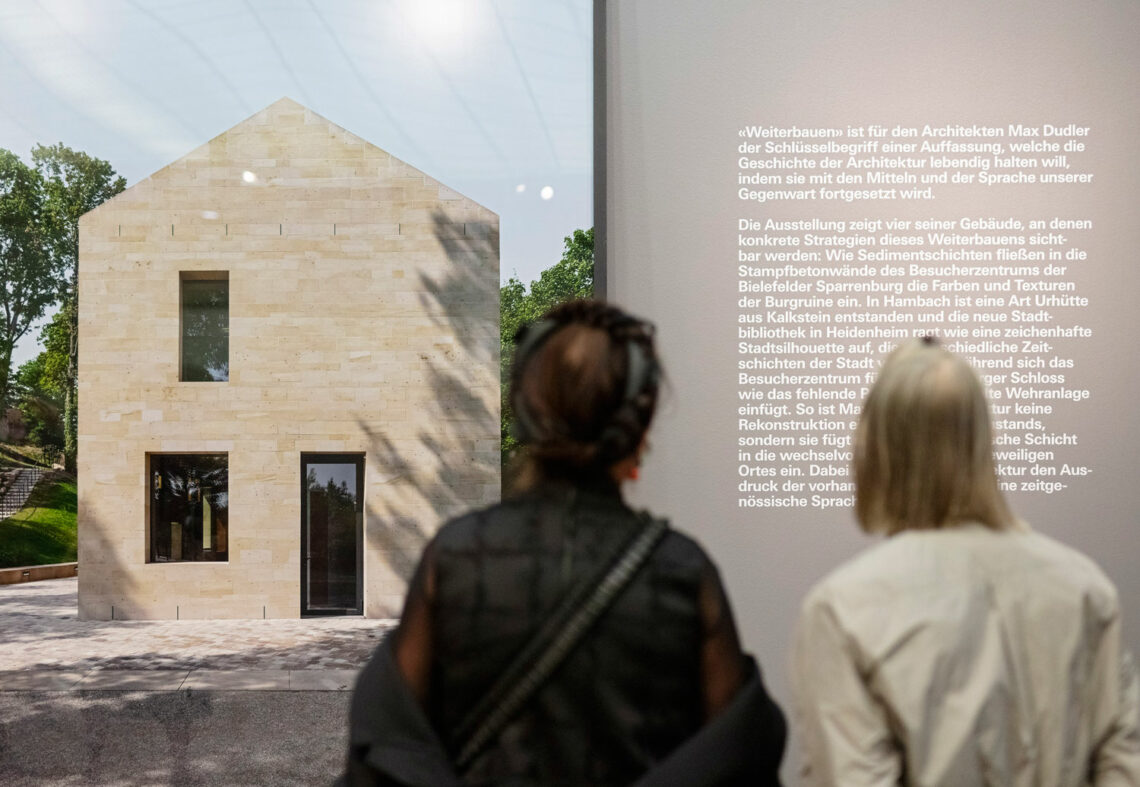 Two people with longer hair from behind. They are looking at a large photo showing a modern house with a friendly stone color and very simple shapes. To the right is a wall text about the exhibition.