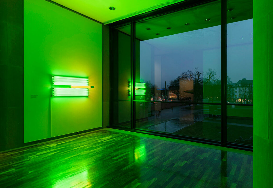 An exhibition room in the Kunsthalle as darkness falls. The room is bathed in greenish-yellow light emanating from a fluorescent tube sculpture on the wall. Outside, trees, houses, lights and the water basin in the park.