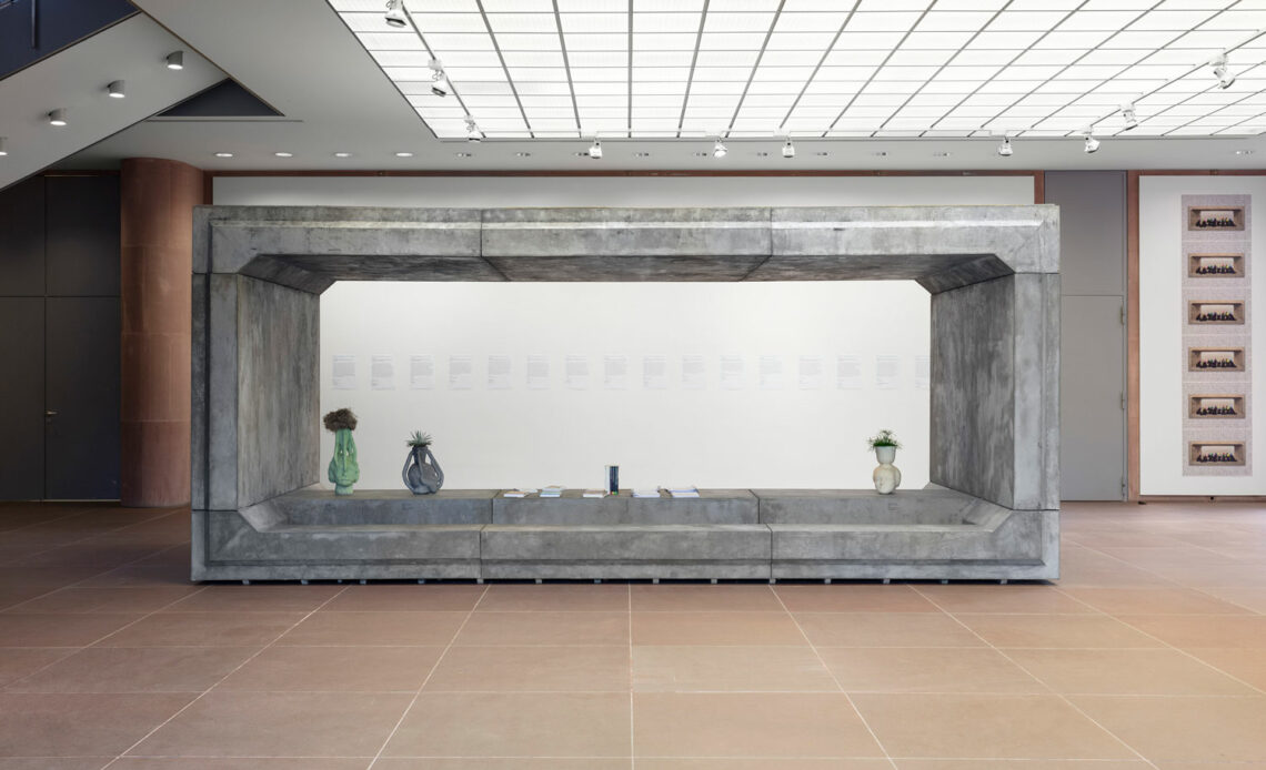 Wie ein Container steht eine Betonstruktur in der Eingangshalle der Kunsthalle. Die beiden langen Seiten sind offen, wir schauen frontal drauf. Darin drei Vasen auf einer Betonfläche und mehrere flache weiße Objekte, vermutlich Bücher.