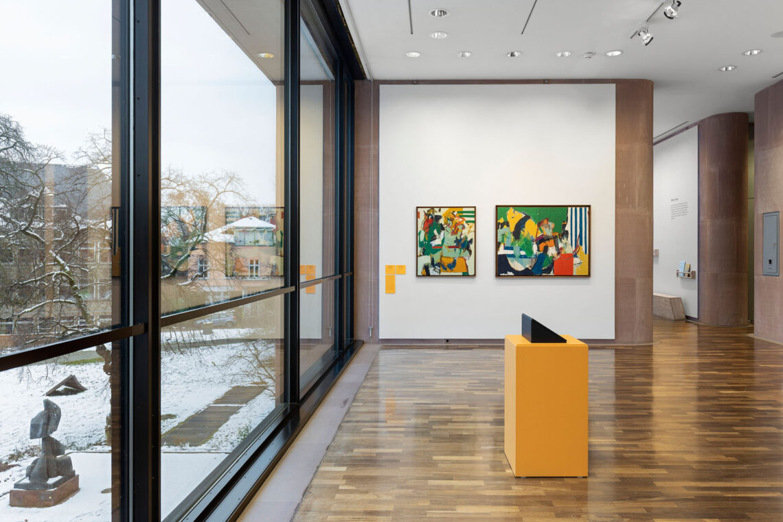 Ausstellungsraum mit Blick in den Skulpturenpark der Kunsthalle. Im Schnee sind zwei Skulpturen. Im Raum ein oranger Sockel mit Monitor, hinten an der Wand zwei Gemälde in unruhigen Grün-, Gelb-, und Blautönen mit etwas Weiß und jeweils ein paar Streifen.