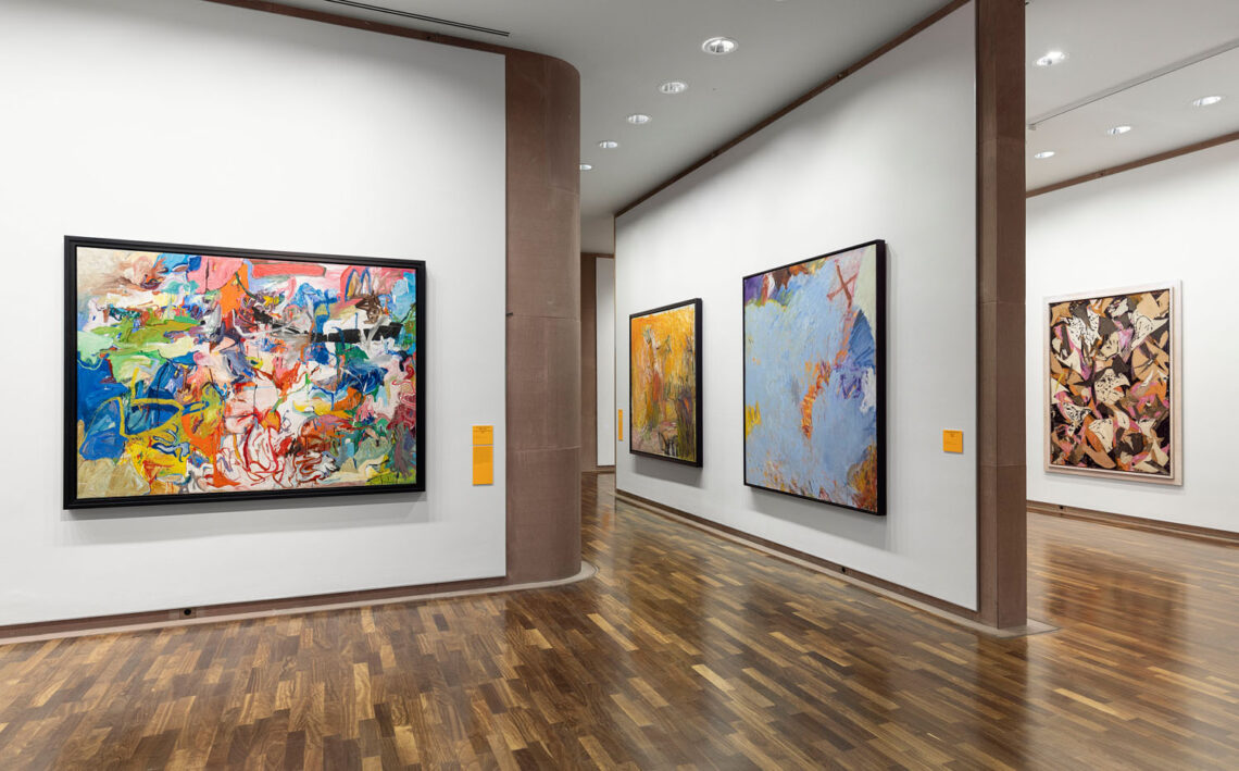 View of three walls in the Kunsthalle. On the left, a very colorful, wild horizontal format with streaks of color, then two square canvases with a more yellow motif and a light blue one. Behind it is a work with triangular shapes in ochre, brown, orange and white.