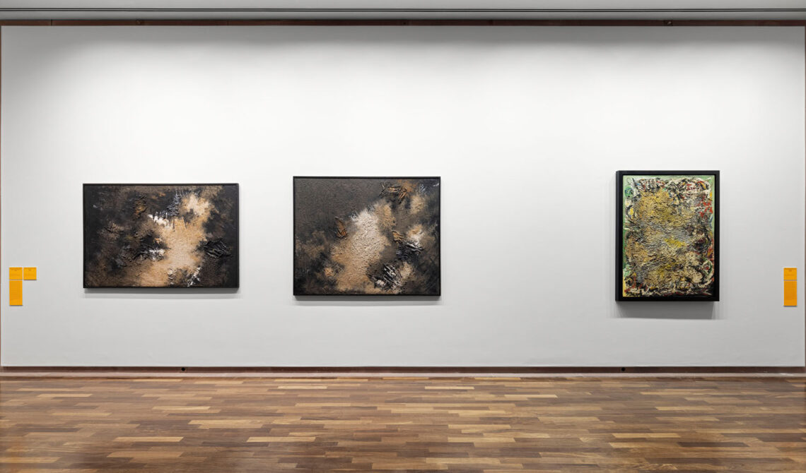 A white wall with three paintings. On the left, two very similar ones with a delicate white structure surrounded by a dark brown surface. On the right, at a distance, a vertical format with more yellowish-green splashes of color.