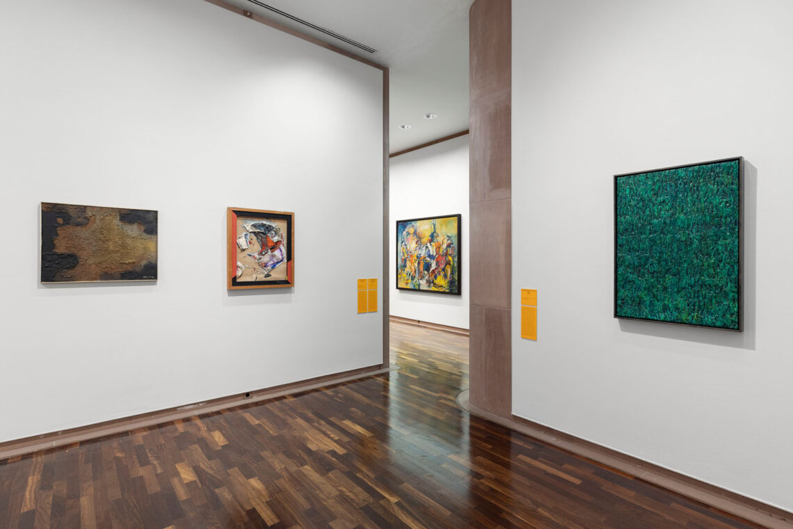 A passageway. On the right wall, a square painting in dark green. Two smaller ones on the left. The one on the left in earth tones, the one on the right with a collage of materials and a handmade wooden frame. In the room behind it, a painting with moving colors, lots of yellow, many splashes of other colors.