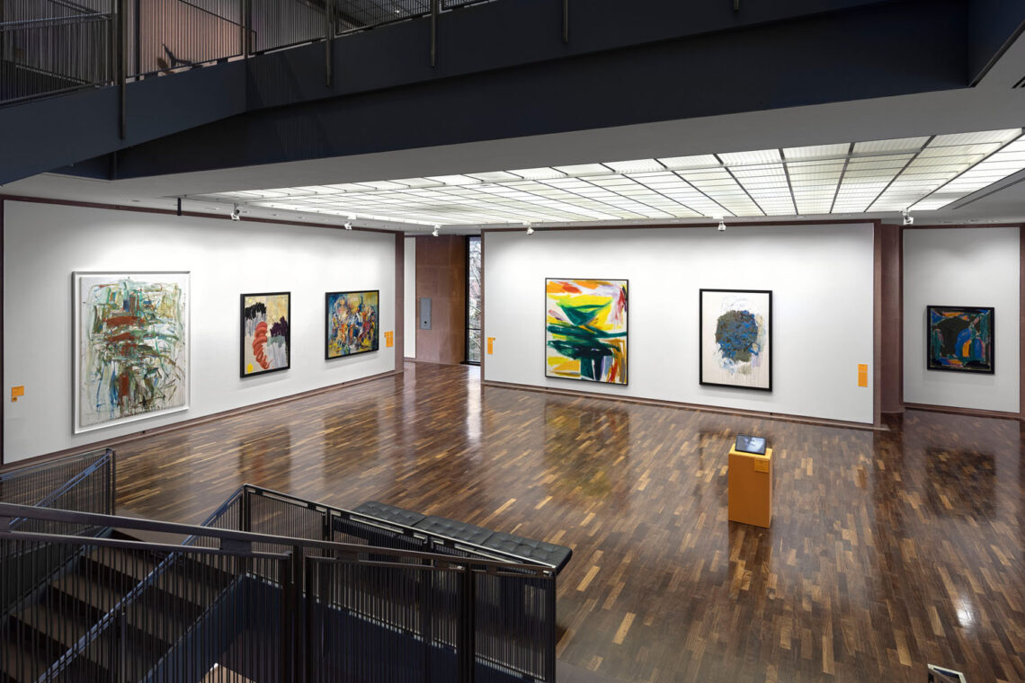 View from the staircase into the main room on the second floor of the Kunsthalle. Shiny parquet flooring, an orange plinth with a monitor in the middle of the room. Large-format abstract paintings on the walls, painted quite dynamically.