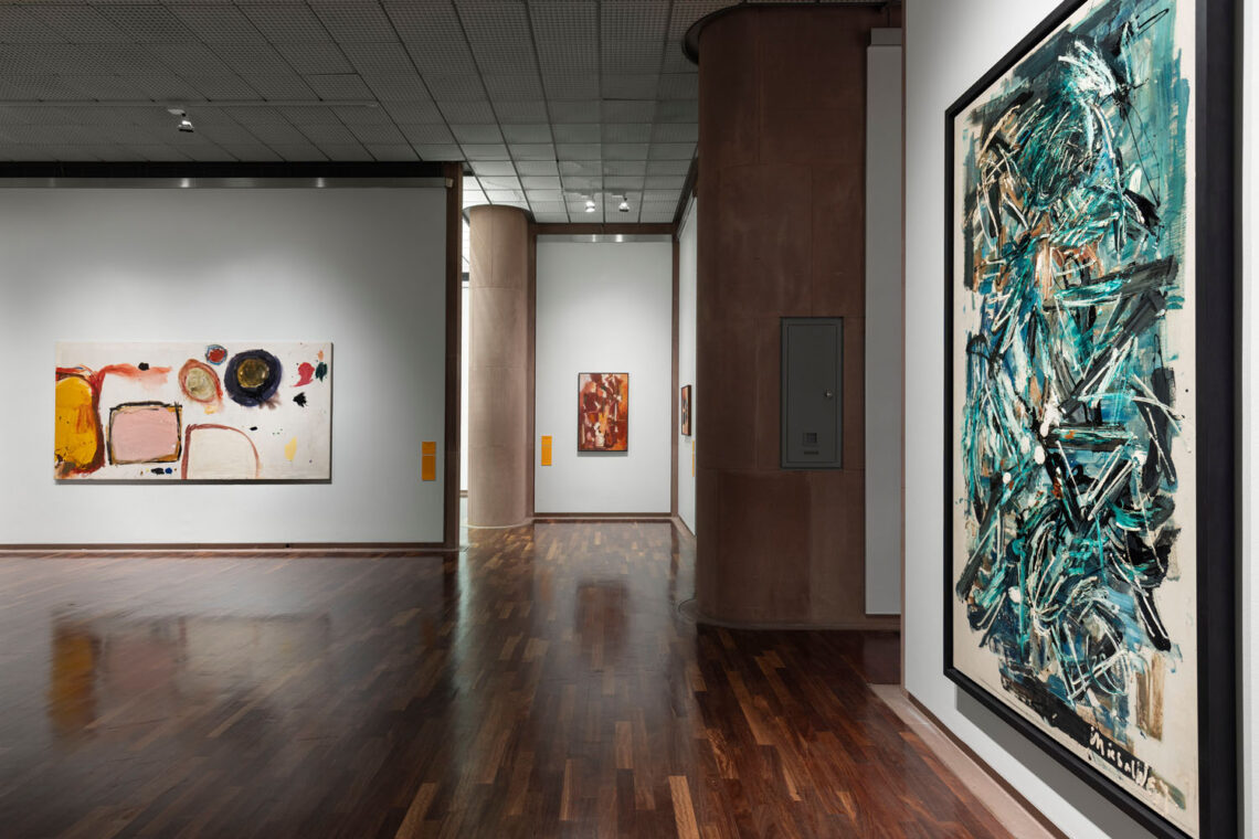 Atmospherically lit exhibition room of the Kunsthalle. On the right wall, a large painting created by rapid movements with a brush. Green and black. On the left wall, a landscape format with simple shapes. At the back, a portrait format in orange and red.