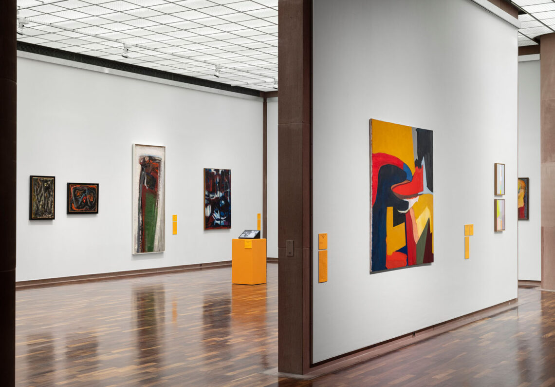 View into two rooms of the Kunsthalle with colorful paintings. In the background, an orange pedestal with a monitor.