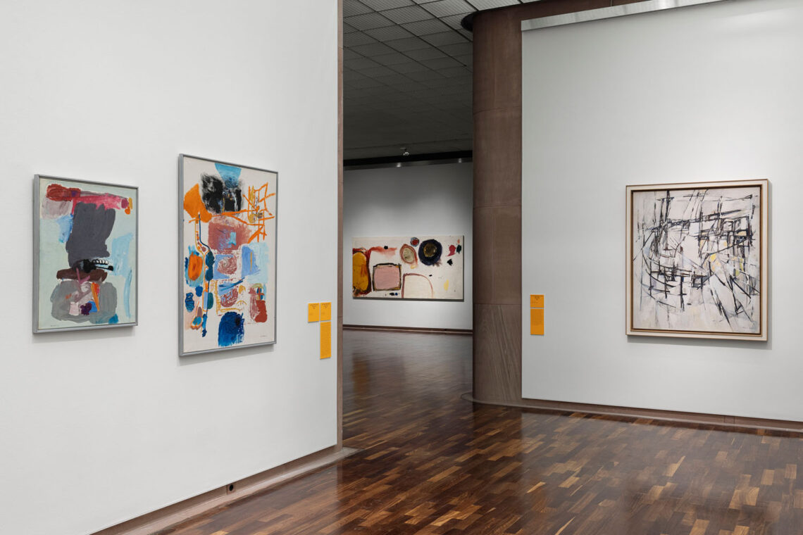 View from one exhibition room into another. Two colorful abstract paintings with shades of grey, blue and orange on the left wall in front. On the right wall, a delicate grid structure in black on a white background. In the distance, a rather large landscape format with simple shapes.