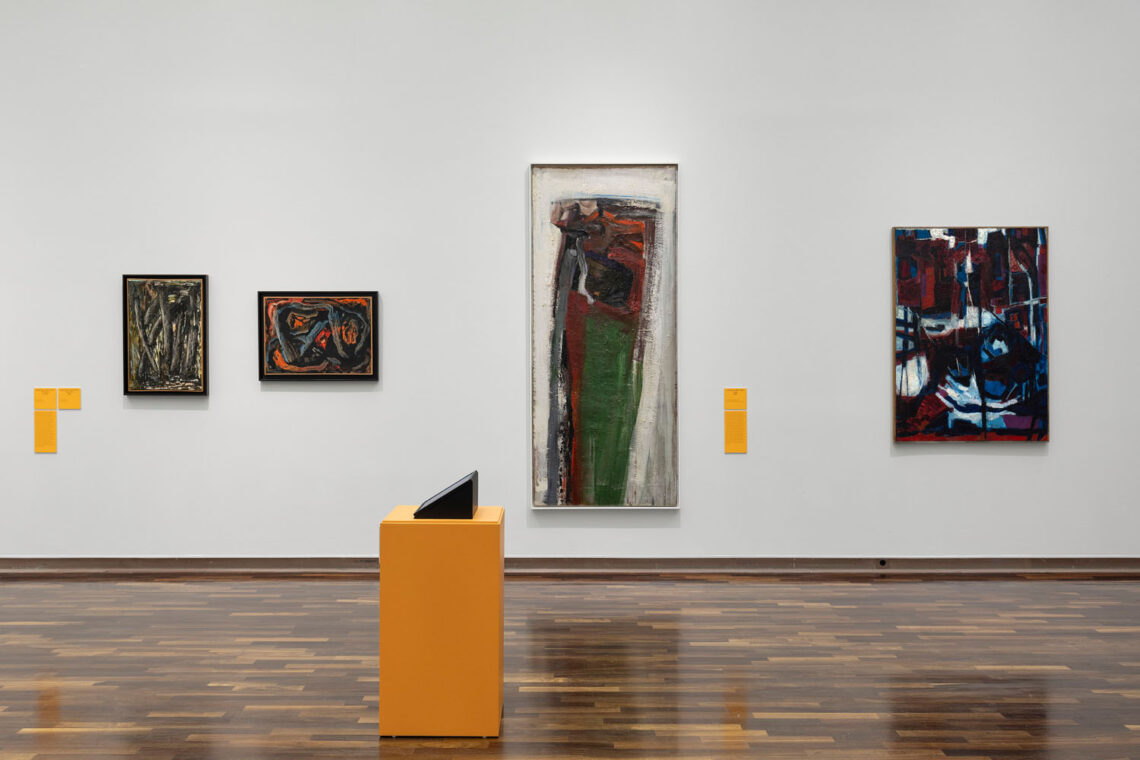 Exhibition room. In front an orange pedestal with a monitor, behind on the wall four paintings of different sizes with quite a large proportion of black. Mostly connected shapes painted with a thick brush.