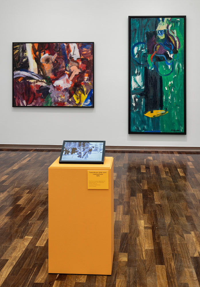 Exhibition room of the Kunsthalle with plenty of parquet flooring. In the foreground is an orange plinth with a monitor showing two people in black and white, holding on to one arm and balancing. Behind them on the wall to the left is a landscape format with wild swirls of color, mainly in shades of red and black. To the right is an extreme portrait format in shades of green, looking a bit like a Big I with a hat.