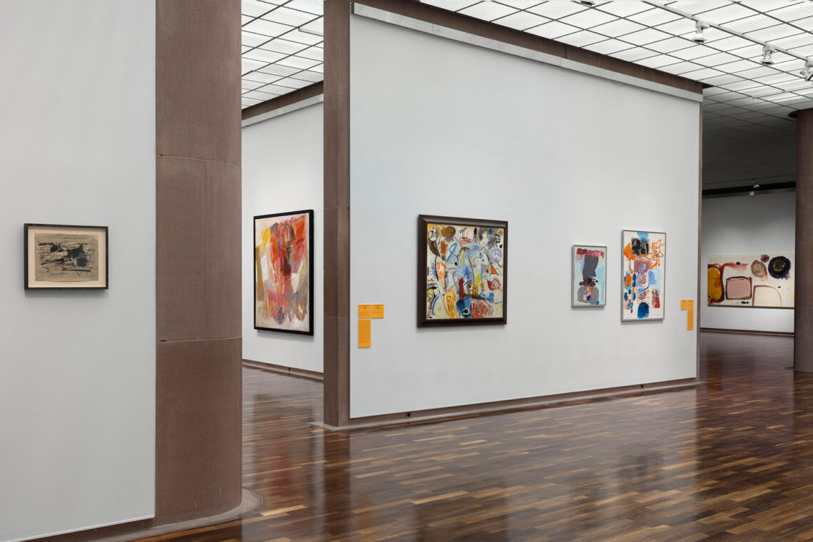 View of several exhibition rooms in the Kunsthalle. On the left of the wall, two smaller paintings or drawings with wildly painted shapes in dark blue or grey colors and a few blue and ochre shapes against a light background. To the right of the wall, a view into two other rooms one behind the other, where there is an orange pedestal with a small black monitor on it, and behind it a very large painting with a few poured areas of color. At the very back and quite small, another painting with thick black lines and lots of light gray is visible.