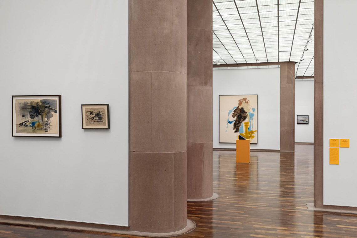 View of several exhibition rooms in the Kunsthalle. On the left wall are two small paintings or drawings with wildly painted shapes in dark blue or grey colors and a few blue and ochre shapes against a light background. To the right of the wall, a view into two other rooms one behind the other, where there is an orange pedestal with a small black monitor on it, and behind it a very large painting with a few poured areas of color. At the very back and quite small, another painting with thick black lines and lots of light gray is visible.