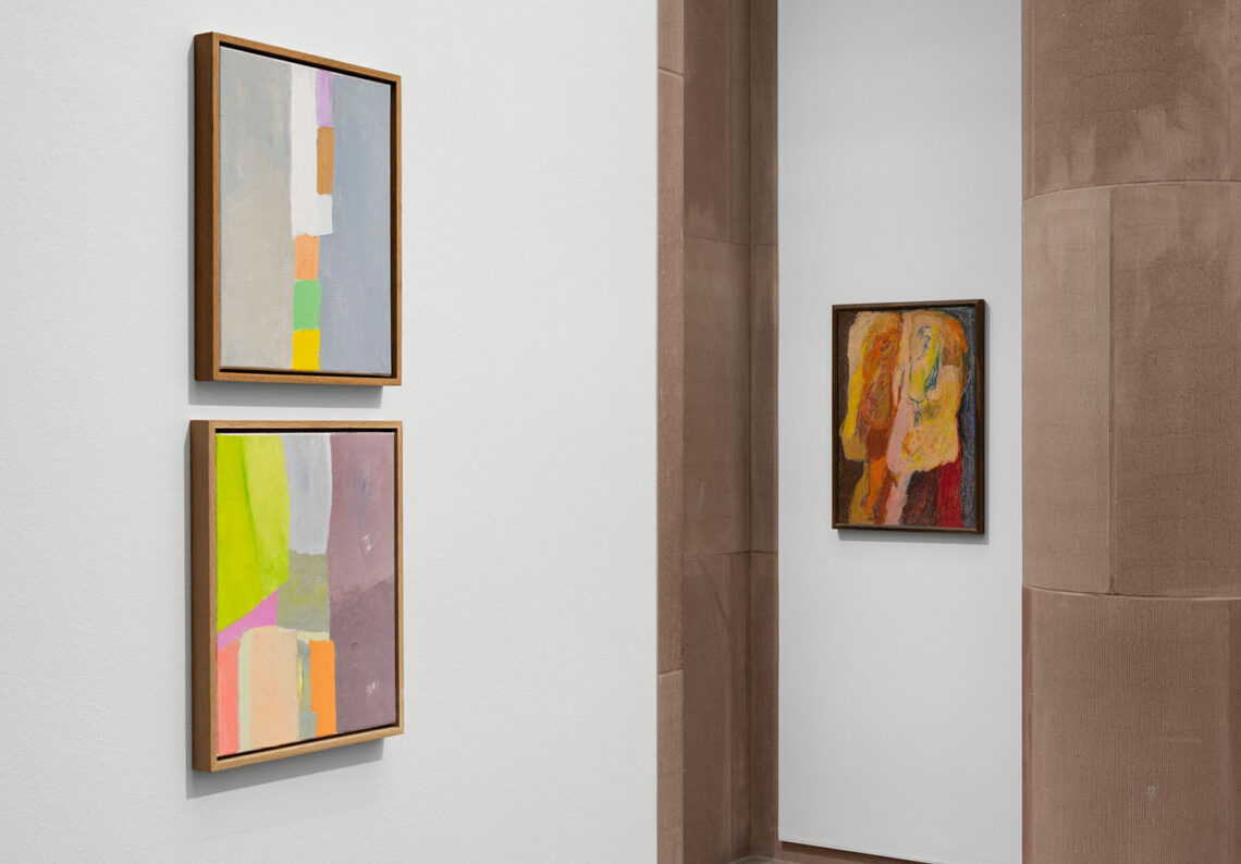 View of two walls. On the left, two small paintings on top of each other with lots of grey, but also cheerful, bright, more angular areas of color. On the other wall, a portrait format with two upright abstract creatures in warm shades of orange, red and ochre against a dark background.
