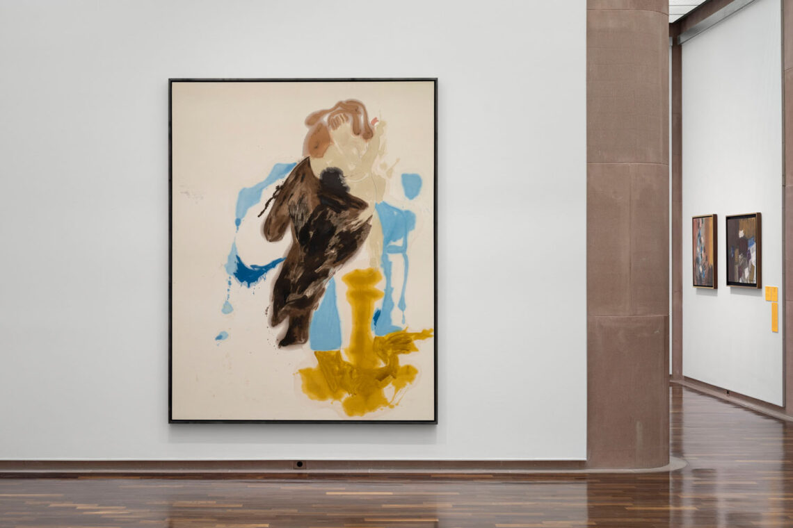 A rather large portrait format hangs on a white wall. Paint has been poured onto the unprimed canvas. The resulting brown, ochre and light blue areas create the impression of a standing female figure. On a wall further back are two other smaller works in darker colors.