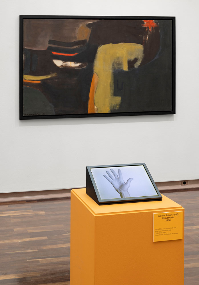 In the foreground, an orange plinth with a slanted monitor showing a hand reaching upwards. Behind it on the wall is a painting in landscape format with a very dark color and only a few areas of color in ochre, orange, red and white. It is slightly reminiscent of an animal head, perhaps a pig from a comic strip.