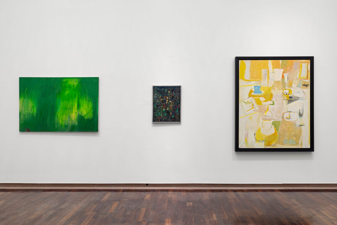 Three abstract paintings hang on a white wall. On the left is a landscape format in shades of green, in the middle a small dark portrait format with splashes of color, on the right a larger portrait format in bright shades of white and yellow that appears to show a table with glasses.