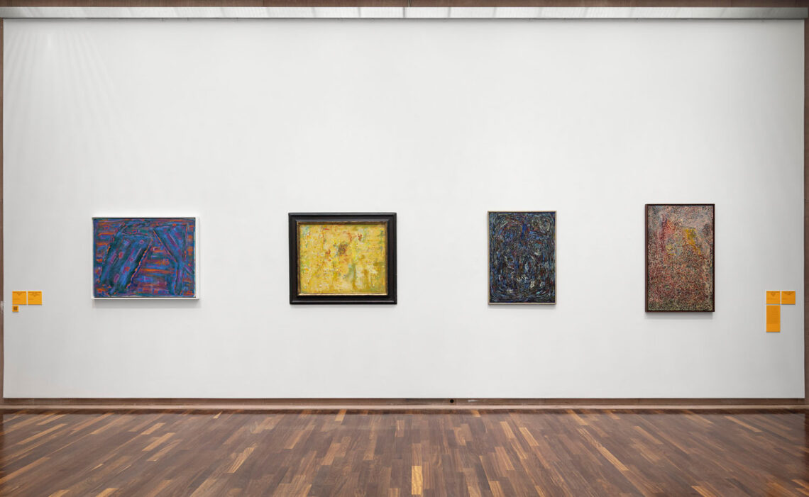 Four paintings on a white wall. On the left a lot of blue and some red, next to it more yellow, then a darker one in shades of blue, on the far right a light painting in darker shades of red, green and orange that looks as if it has been spotted.
